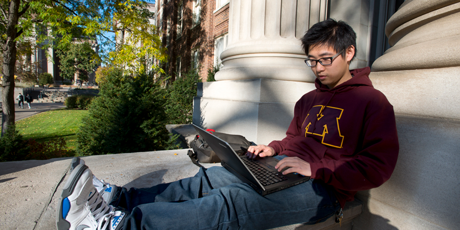 A student works on his laptop while sitting against a pillar outside